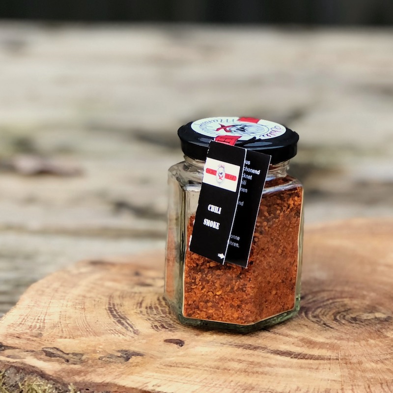 geräuchertes Chili Pulver im Glas con der Chilli Manufaktur auf einer Holzscheibe auf einem Tisch stehend